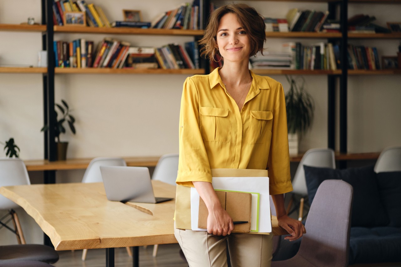 Young woman in yellow shirt leaning on desk with notepad and papers in hand happily looking in camera in modern office
